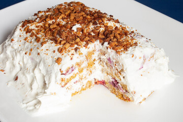 Close-up of a homemade dessert with a slice taken out, topped with whipped cream and sprinkled with crunchy nut pieces, served on a pristine white plate that showcases its creamy layered interior.