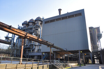 Naklejka premium The photograph depicts an old metallurgical plant in Duisburg. Rusty metal structures and pipes dominate the foreground, highlighting the industrial heritage of heavy steel production. The factory's c