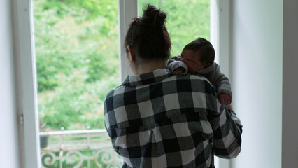 Mother standing by a window with her newborn baby on her shoulder, capturing a serene moment of...