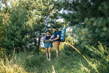 Happy couple tourists with backpacks looking at the map for searching right direction, walking outdoors in the summer forest