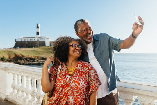 Morden Retirement: Elderly couple taking a selfie at the seaside with a lighthouse in the background - Powered by Adobe