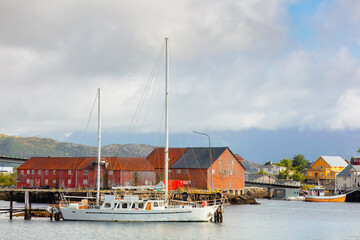 Charming view of Henningsv&aelig;r harbor, with boats docked by rustic old buildings and vibrant red and yellow houses. Misty mountains and a cloudy sky. Henningsv&aelig;r, Norway