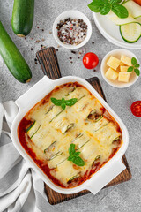 Baked zucchini rolls with ground meat, tomato sauce and cheese in a white baking dish on a gray concrete background. Selective focus, top view.