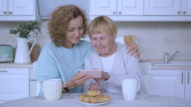 Grandmother and granddaughter sit at kitchen table with cups of coffee and delicious pastries, granddaughter teaching grandmother how to use mobile phone strong family bond love between generations.