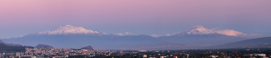 Mexico City nestled at the foot of two snow-capped volcanoes under a soft pink twilight sky.