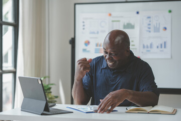 A man celebrating success during an online business meeting with laptop, documents, and charts in the background in an office setting.