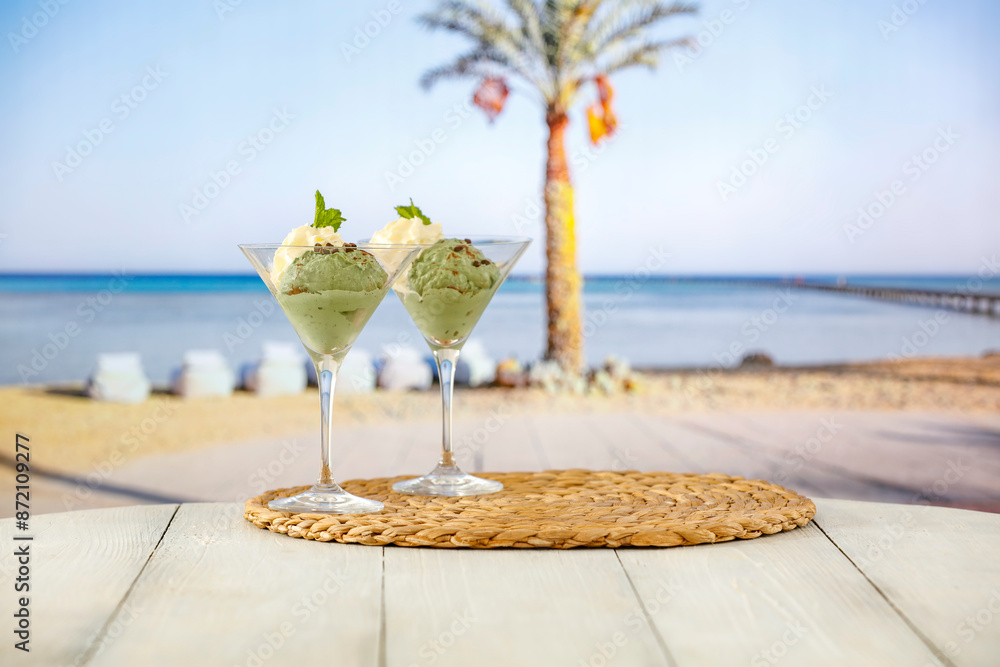 Poster Two glass cups of vanilla and mint ice cream on wooden board table on sandy tropical beach 