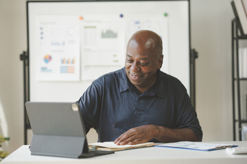 Smiling businessman working on a tablet at his desk, reviewing documents and charts, demonstrating a productive and positive work environment.