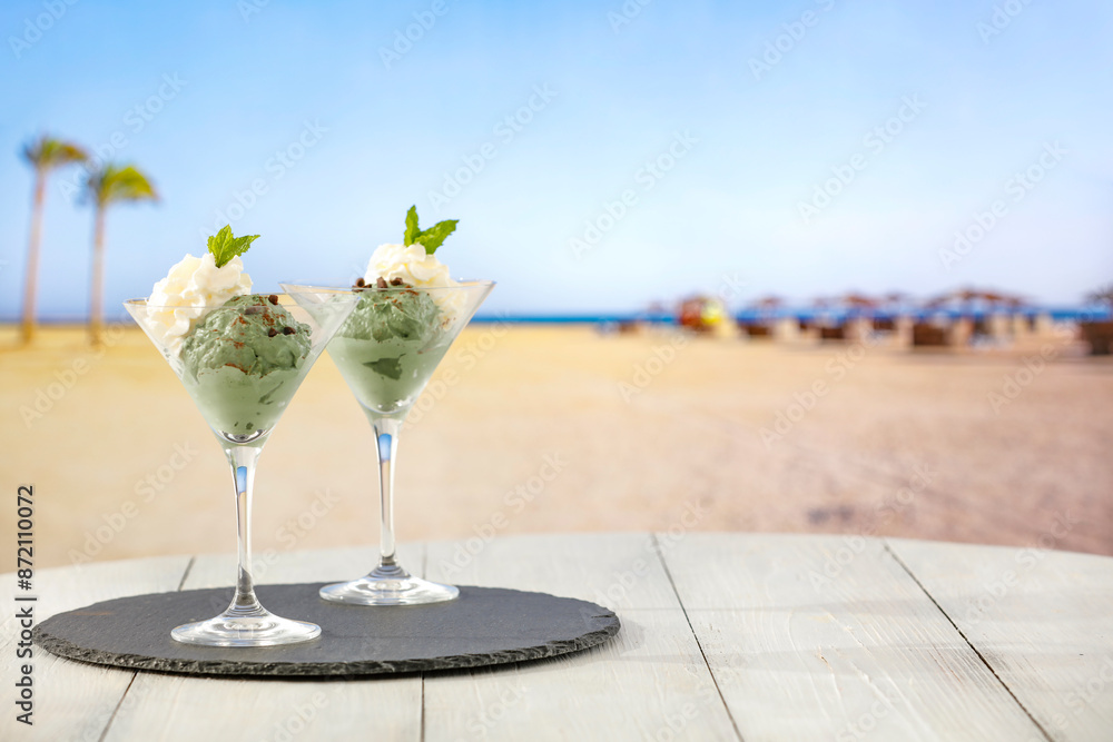 Wall mural Two glass cups of vanilla and mint ice cream on wooden board table on sandy tropical beach 