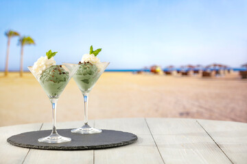 Two glass cups of vanilla and mint ice cream on wooden board table on sandy tropical beach 