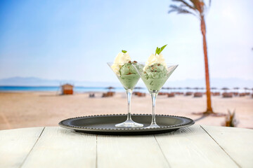 Two glass cups of vanilla and mint ice cream on wooden board table on sandy tropical beach 