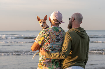 Fototapeta premium homosexual couple back to back with their dog on their lap looking at the camera, on the beach