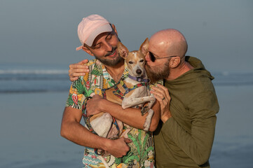 portrait of a couple of gay couple with their dog on their lap, kissing the pet, on the beach at sunset