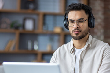 Close-up photo of a serious and focused young Indian man wearing headphones working and studying on a laptop at home