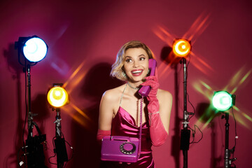 Woman in pink dress and gloves smiles on vintage phone amidst stage lights.