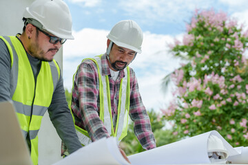 Asian male engineers in safety vests and hard hats reviewing blueprints on outdoor construction...