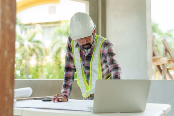 Asian male engineer in safety vest and hard hat reviewing blueprints on table at indoor construction site. Laptop, phone, and architectural plans. Professional, focused on planning and project details