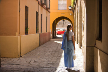 Young beautiful woman with brown hair, sunglasses and blue jacket suit, posing with her back turned under the sun rays on a lonely street. Concept beauty, fashion, trend.