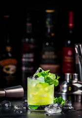 Green Negroni cocktail drink with gin, aperitif, liqueur, mint and ice. Black bar counter background with bottles