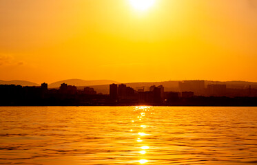 Sea panorama of the city at sunset, silhouettes of the coast and buildings against the backdrop of the orange setting sun reflected in the water