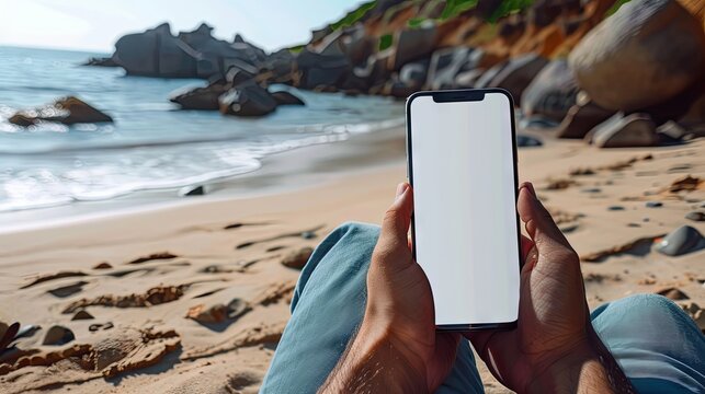 Mockup image of a man holding mobile phone with blank desktop screen while sitting on the beach