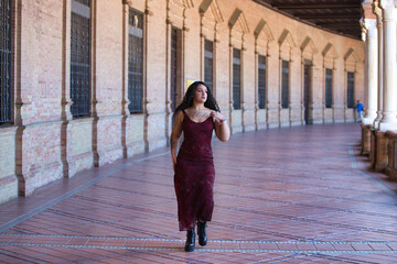 Latin woman, young, beautiful and fat with curves and long curly brown hair, with an elegant long dress walks between the arches of the plaza espana in seville doing different poses and postures.