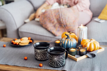 Cozy Halloween plans at home. Hot tea drink in black mugs, cookies and sweets on the plate, pumpkin decor on the coffee table with blurred relaxing woman. Festive autumn, fall mood home time.