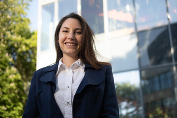 Portrait of a happy caucasian brunette woman in a blue suit stands against the background of a business building in the park and smiles