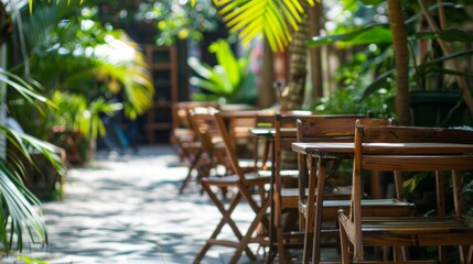 A row of empty wooden tables and chairs sit in a sunlit courtyard, surrounded by lush greenery. The natural light casts shadows on the stone ground, creating a serene and inviting atmosphere