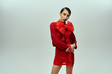 A young woman with short hair poses in a red dress with a large bow. The background is a neutral grey.