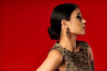 A young woman poses against a bright red background wearing a leopard print top and gold earrings.