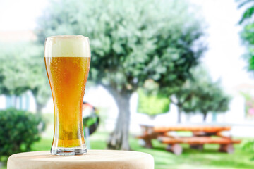 Cold beer with foam  in large glass on wooden table top with green trees in garden in holiday resort background. 