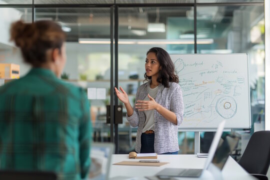 Asian woman giving a presentation to a colleague in a modern office with a whiteboard. Concept of teamwork, communication, and leadership, ideal for coworking day - Powered by Adobe