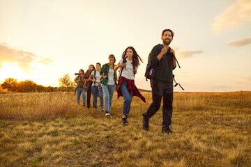 Group of young friends tourists trekking with backpacks walking in a row in the field. Happy people men and women on a hiking trip holiday in nature at sunset. Adventure, travel and tourism concept.