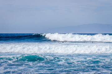 Fototapeta premium Waves coming ashore on a sunny day and surfers ready to surf the waves. Wall of blue wave with white foam coming ashore where surfers are surfing. Atlantic Ocean. Tenerife, Canary Islands, Spain.