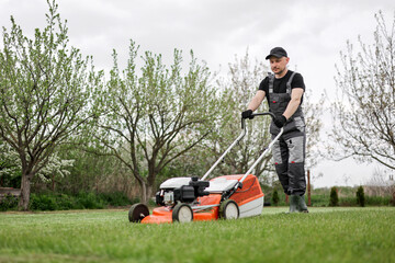 Professional gardener in protective apparel is mowing green grass lawn using modern gasoline...