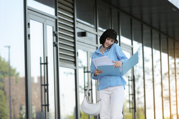 Fototapeta premium Multitasking busy young business woman communicates by smartphone during walks reading and dropping important finance documents by the office buildings outdoors. Stress resistance and business concept
