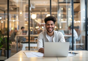 Obraz premium happy young man is sitting at his desk in an office, with a laptop and documents on the table