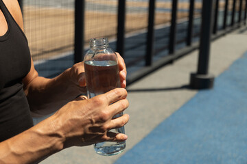 Athletes man holding glass bottle of water on outdoor sports ground. Concept water balance for hot season for active workout. Close up. Copy space.