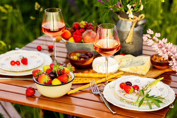 Romantic table decor for a loving couple on blooming meadow with purple lupines. Two glasses of wine, flowers in a vase, silverware, fruits, wooden furniture. Sunset, golden hour, atmospheric light
