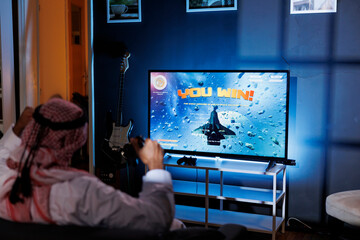 A young Arab man plays an immersive virtual reality game at home, celebrating victory while holding...