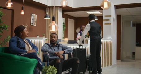 Naklejka premium Woman wheelchair user sitting in lounge area and waiting for accommodation check in with husband, dealing with physical disability and travelling on honeymoon. Chronic impairment.