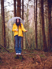 Teenager girl in yellow jacket, blue jeans and translucent umbrella standing on a tree trunk checking out rain. Trip to a forest park. Explore nature theme. Selective focus.