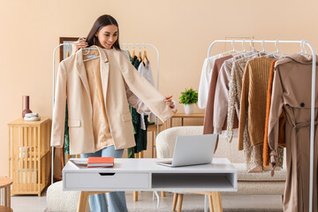 Happy female stylist with laptop and clothes video chatting on rack in studio