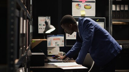 African American detective inspects evidence in well-organized office, focused on solving crime. Dedicated police officer approaching his desk while holding case folder for criminal investigation.