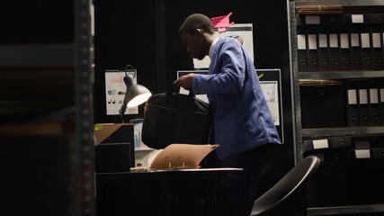 While holding a laptop bag, the private detective stands and goes out of the office. African american police officer closes his laptop and prepares his workstation before departing for the day.