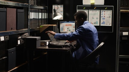 African American detective inspector arrives at office, investigating crime. Clues, evidence, files, photos, and records fill the room. Laptop bag indicates reliance on technology.