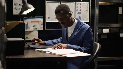 Private investigator seen in dimly lit office meticulously analyzing papers and evidence gathered during research. With laptop open, policeman cross-references information and does background check.