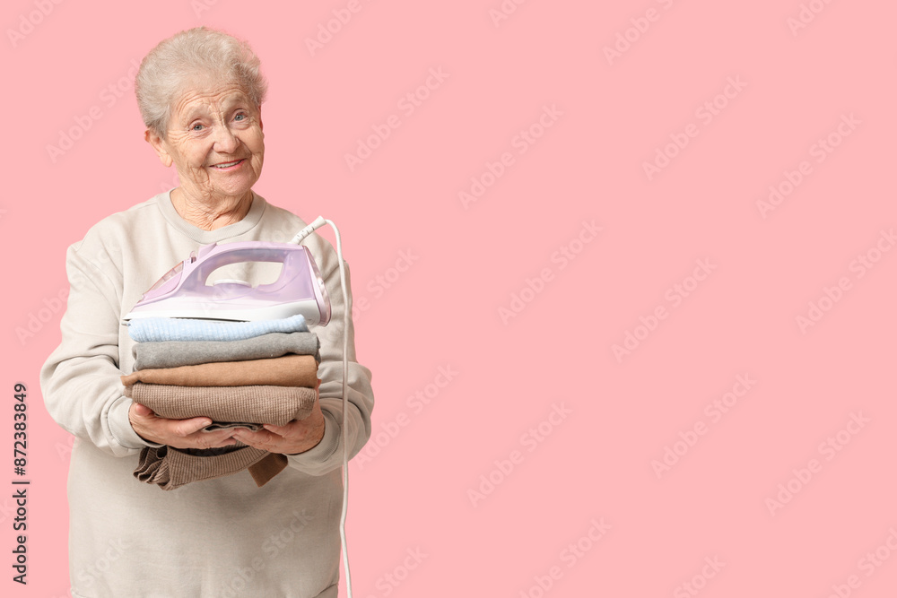 Wall mural Happy senior woman with iron and stack of laundry on pink background