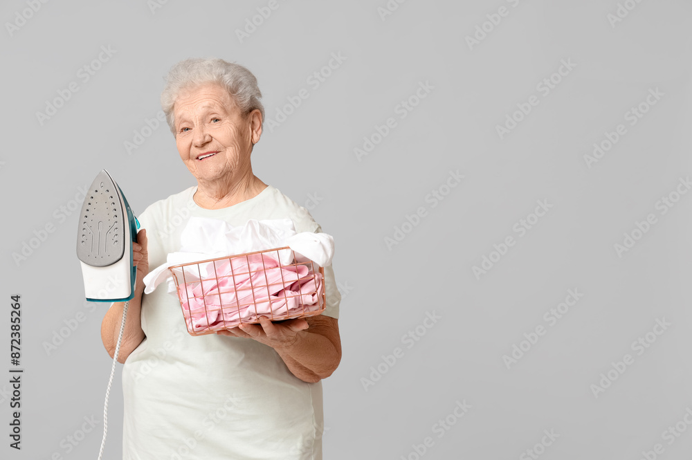 Wall mural Happy senior woman with iron and basket of laundry on grey background